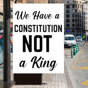 Puede incluir: Un letrero blanco con texto negro que dice "We Have a CONSTITUTION NOT a King." El letrero se muestra al aire libre, con una calle de la ciudad y edificios al fondo.