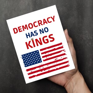Puede incluir: Una tarjeta blanca con el texto "DEMOCRACY HAS NO KINGS" en rojo y azul. Debajo, un dise&ntilde;o de bandera estadounidense desgastada en rojo y azul. La tarjeta se sostiene en una mano.