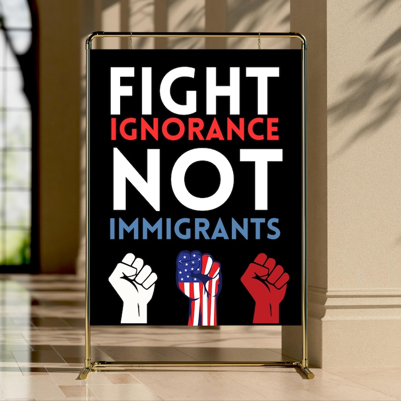 May include: A black sign with the words "FIGHT IGNORANCE NOT IMMIGRANTS" in white, red, and blue. The sign features three fists: white, American flag design, and red. The sign is displayed on a gold metal stand.