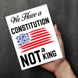 May include: A white card with the text "We Have a CONSTITUTION NOT a KING" in black script and bold font. The card features a stylized American flag with red stripes and blue stars.