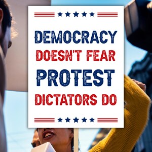 May include: A white sign with the words "DEMOCRACY DOESN'T FEAR PROTEST DICTATORS DO" in blue and red, with stars and stripes design. The sign is held up in a crowd.