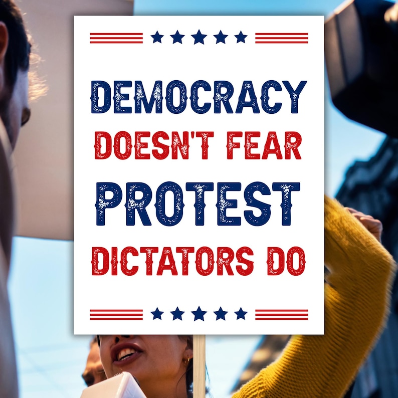 May include: A white sign with the words "DEMOCRACY DOESN'T FEAR PROTEST DICTATORS DO" in blue and red, with stars and stripes design. The sign is held up in a crowd.