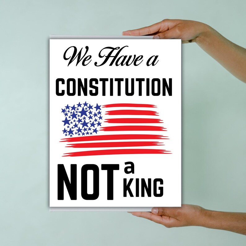 May include: A white rectangular sign with the text "We Have a Constitution NOT a King." The word "Constitution" is in bold, large, black letters. The American flag is depicted with blue stars and red stripes.