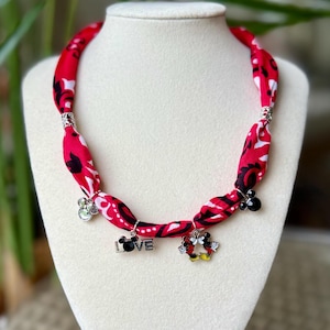 May include: Red fabric necklace with black and white floral pattern. The necklace has silver accents and charms with the word "LOVE" and Mickey Mouse designs. The necklace is displayed on a white mannequin.