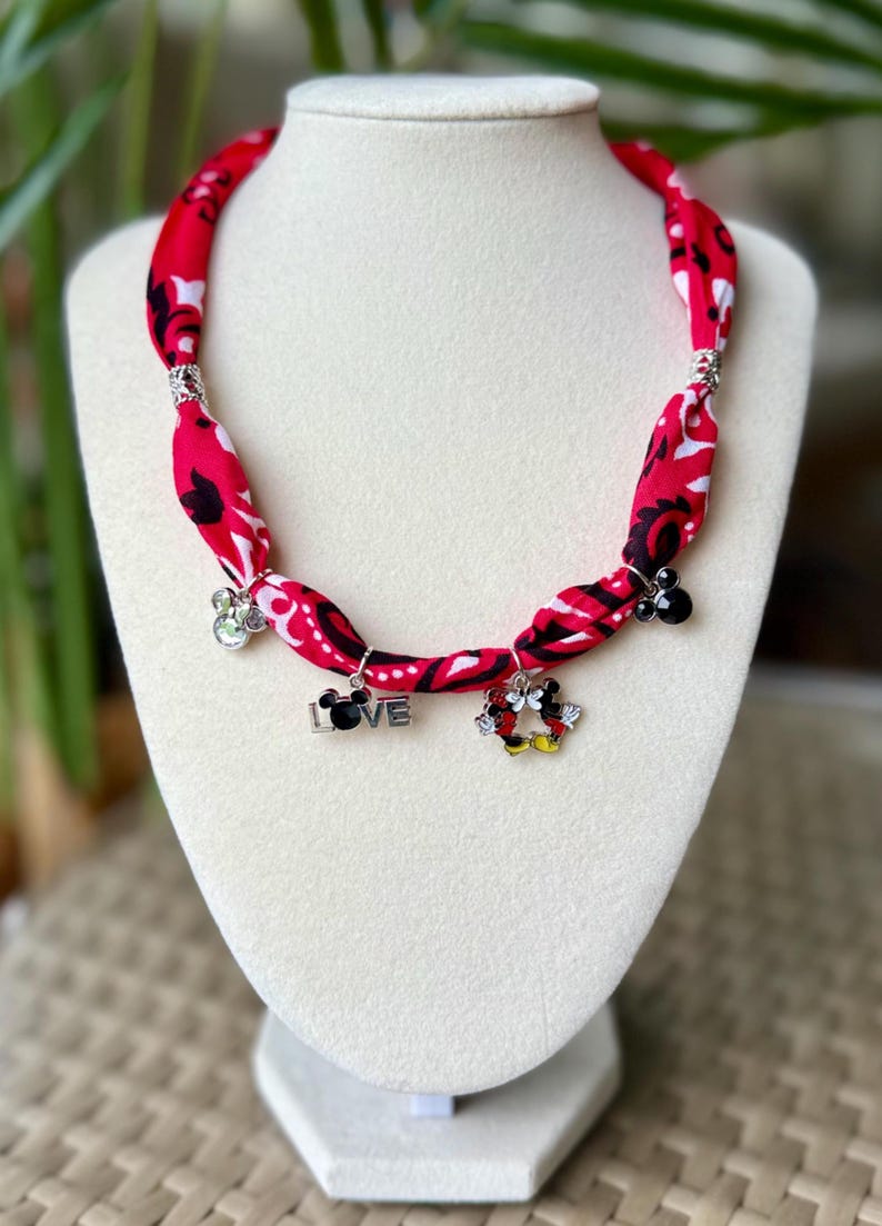May include: Red fabric necklace with black and white floral pattern. The necklace has silver accents and charms with the word "LOVE" and Mickey Mouse designs. The necklace is displayed on a white mannequin.
