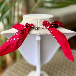 May include: A red bandana necklace with white polka dots and black paisley designs. The necklace is displayed on a white mannequin bust and features a silver-colored clasp. The ends of the bandana are tied into bows.