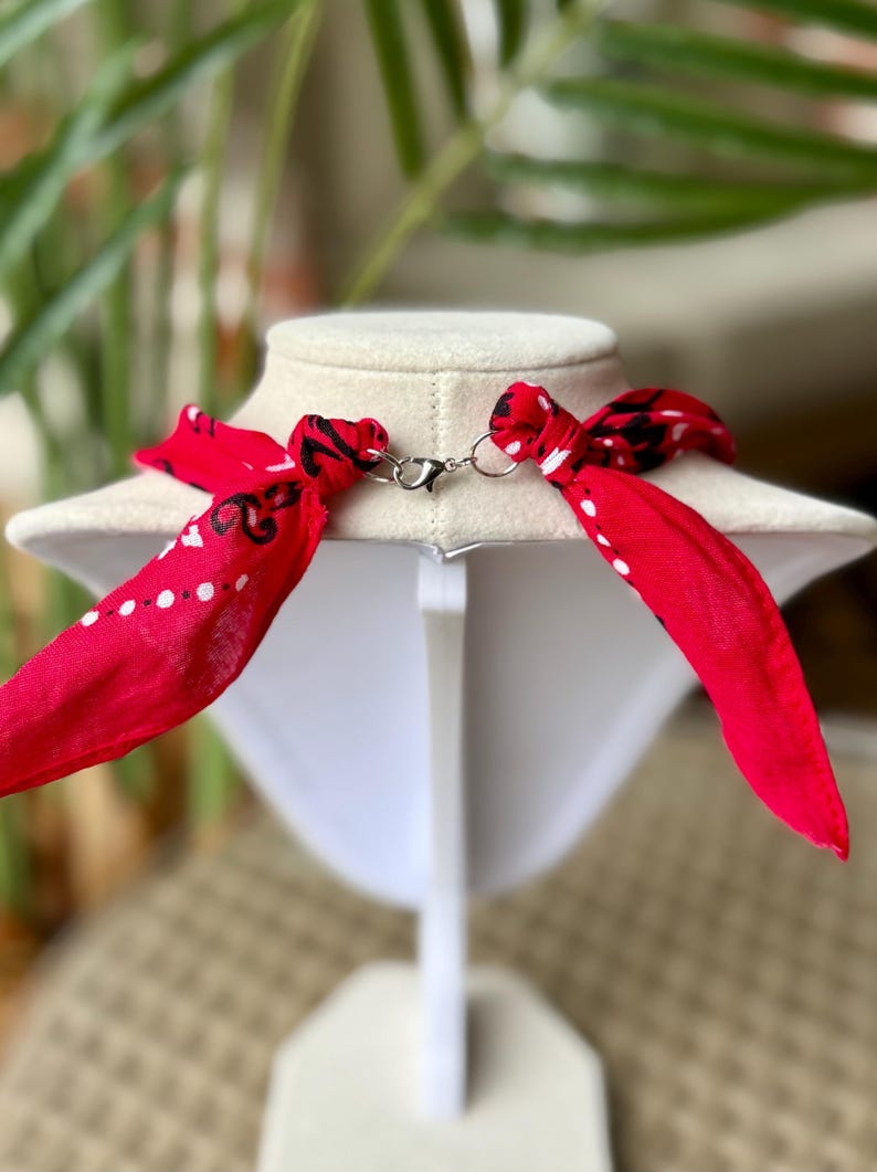 May include: A red bandana necklace with white polka dots and black paisley designs. The necklace is displayed on a white mannequin bust and features a silver-colored clasp. The ends of the bandana are tied into bows.