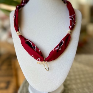May include: A red fabric necklace with a white and black pattern, adorned with gold-colored metal accents and a Florida state charm. The necklace is displayed on a white mannequin head, showcasing its design and style.