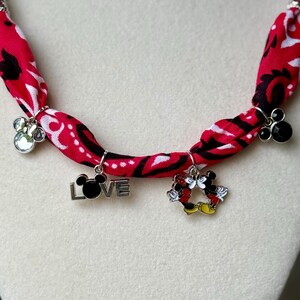 May include: A red fabric necklace with a black and white pattern. The necklace features silver charms, including a paw print, the word "LOVE" with a Mickey Mouse head, and a Mickey and Minnie Mouse design.