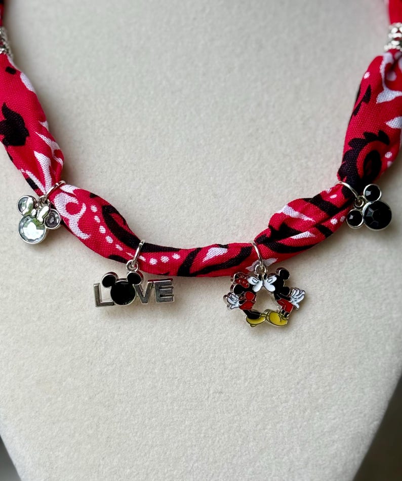 May include: A red fabric necklace with a black and white pattern. The necklace features silver charms, including a paw print, the word "LOVE" with a Mickey Mouse head, and a Mickey and Minnie Mouse design.