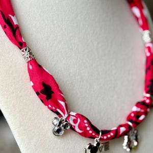 May include: A red fabric necklace with a black and white pattern. The necklace features silver charms with a Mickey Mouse design and the word "LOVE". The necklace is displayed on a white surface.