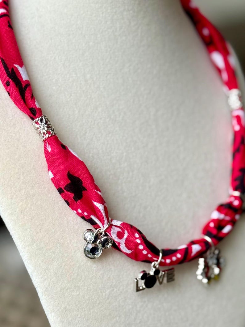 May include: A red fabric necklace with a black and white pattern. The necklace features silver charms with a Mickey Mouse design and the word "LOVE". The necklace is displayed on a white surface.