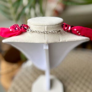 May include: A necklace with a silver chain and a bright pink scarf with white polka dots. The necklace is displayed on a white jewelry bust against a blurred background.