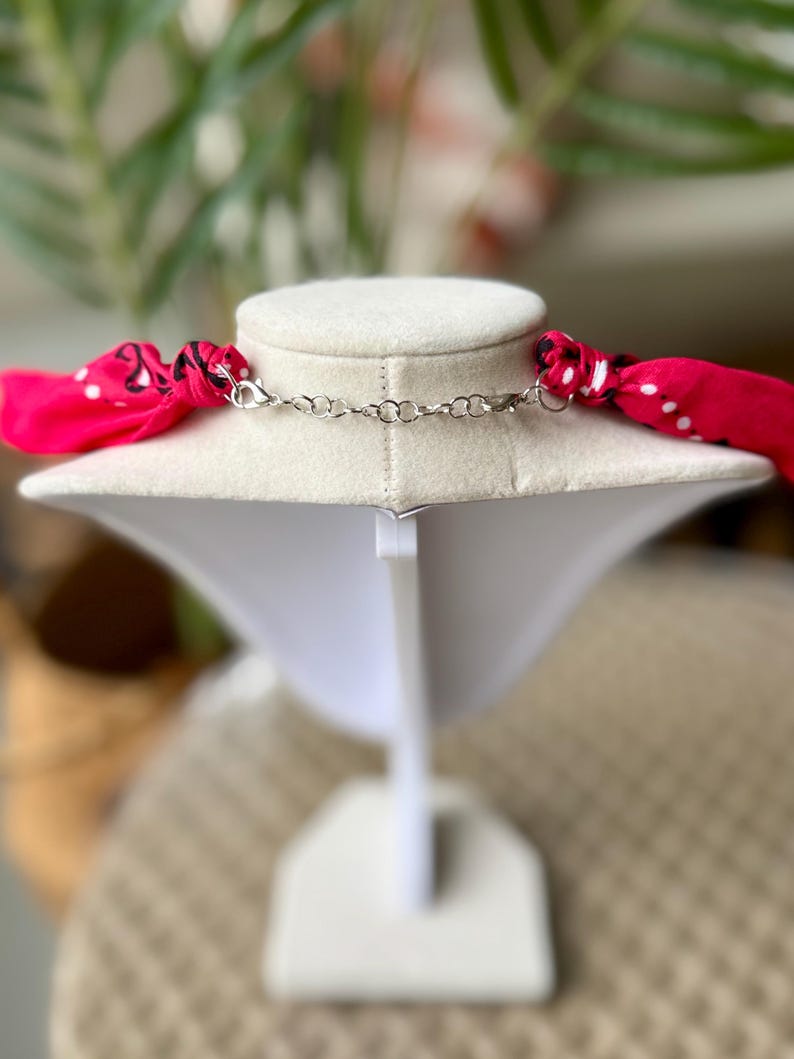 May include: A necklace with a silver chain and a bright pink scarf with white polka dots. The necklace is displayed on a white jewelry bust against a blurred background.