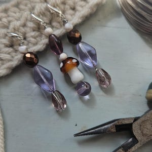 May include: Handmade beaded earrings with a mushroom charm, purple, brown, and clear beads. The earrings are displayed on a cream-colored crocheted surface, with a pair of pliers in the background.