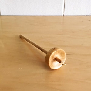 May include: A wooden drop spindle with a long, slender shaft and a circular whorl. The spindle is made of light-colored wood and has a small hook at the bottom of the whorl. The background is a light-colored wooden surface.