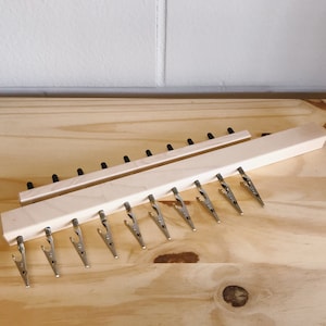 May include: Two wooden display racks with metal clips. One rack has 10 clips, the other has 11. The racks are made of light-colored wood and are designed to hold items. The background is a light-colored wooden surface.