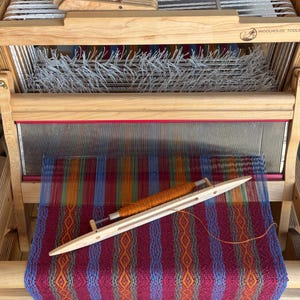 May include: A light-colored wooden table loom with the brand name Woolhouse Tools printed on the frame. The loom features a metal reed, white warp threads, and a woven fabric with a colorful geometric pattern in red, blue, and orange.