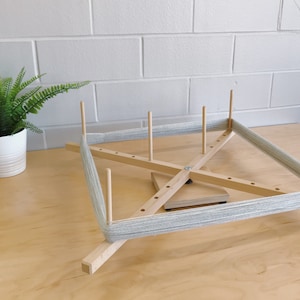 Puede incluir: Un enhebrador de lana de madera con lana gris enrollada alrededor. El enhebrador tiene cuatro brazos de madera y una base central giratoria.