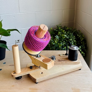 May include: A wooden yarn swift with a vibrant pink, purple, and orange yarn ball. The swift has a natural wood finish and a black motor. The yarn is wound around a wooden spindle.