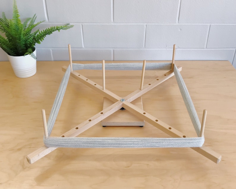 Puede incluir: Un enhebrador de lana de madera con lana gris enrollada alrededor. El enhebrador tiene cuatro brazos y un eje central.