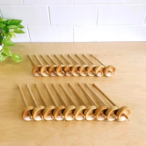 May include: A collection of wooden drop spindles arranged in two rows. Each spindle has a long, slender handle and a circular, disc-shaped whorl. The wood has a light, natural finish.