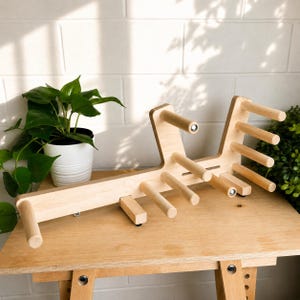 May include: A light-coloured wooden yarn storage rack featuring a horizontal base with multiple vertical pegs extending upwards and outwards. The structure is crafted from smooth, natural wood and includes several cylindrical dowels arranged in a tiered formation.