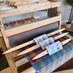 May include: A wooden weaving loom with a colorful woven textile in progress. Three wooden shuttles with "The Surf Shuttle" labels are placed on the woven fabric. The loom is made of light-colored wood.