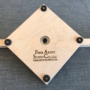 Puede incluir: Una base cuadrada de madera con cuatro patas de goma negra y un rodamiento de metal en el centro. La base est&aacute; etiquetada con "Fiber Artist Supply Co., LLC" y "fiberartistsupply.com".