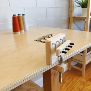 May include: A wooden yarn swift clamp with metal clips and hooks, attached to a light-colored wooden table. Three spools of yarn in orange, gold, and white are in the background. The clamp is designed to hold yarn for crafting.