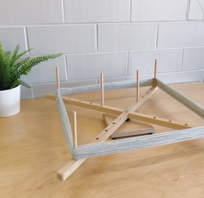 Puede incluir: Un enhebrador de lana de madera con lana gris enrollada alrededor. El enhebrador tiene cuatro brazos de madera y una base central giratoria.