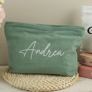May include: A sage green corduroy pouch embroidered with the name "Andrea" in white script. The pouch is rectangular and rests on a woven mat. Another white pouch with black script is visible.