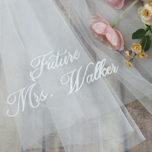 Pode incluir: Um véu de noiva branco transparente com o texto bordado "Future Mrs. Walker" em escrita elegante. O véu é drapeado, com dobras suaves, e é realçado por delicadas flores cor-de-rosa.