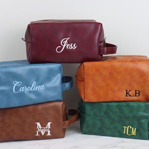 Op de afbeelding: Een stapel van zes gepersonaliseerde toilettassen in verschillende kleuren: bordeaux, blauw, bruin, bruin en groen. Elke tas heeft een handvat en een ritssluiting. De tassen zijn geborduurd met namen of initialen in wit of geel garen.