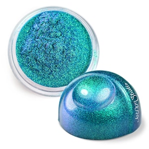 Pode incluir: Um pequeno frasco de pigmento de glitter azul turquesa iridescente. O glitter é solto e tem um acabamento brilhante. O frasco é de plástico transparente e tem um rótulo branco com o texto "Jouer Studio".