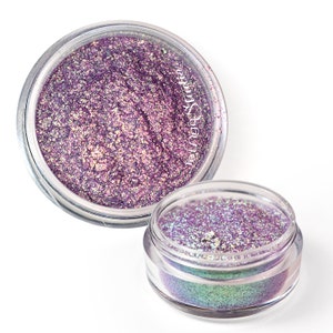 Puede incluir: Dos pequeños frascos de purpurina púrpura iridiscente. La purpurina está en un frasco transparente con una tapa negra. Los frascos están sobre un fondo blanco.