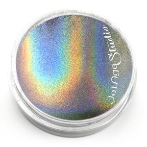 Puede incluir: Un recipiente redondo de plástico transparente lleno de polvo holográfico fino que refleja un espectro de colores arcoíris, incluyendo azul, verde y dorado. El texto Jetnga Studio está impreso en letra cursiva blanca en el lado derecho.