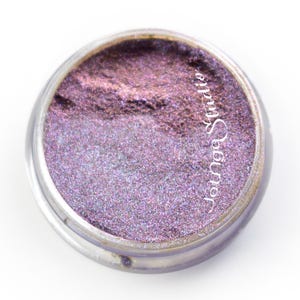 May include: A clear, round jar filled with shimmering purple eyeshadow. The eyeshadow has a gradient effect, transitioning from a darker, more metallic shade to a lighter, iridescent hue. The text "Jotnge Studio" is printed on the side.
