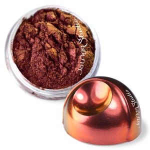May include: A jar of loose, shimmering eyeshadow in shades of burgundy and gold. The open container reveals the product's texture, while the detached lid, with the text "Jotnge Studio", reflects the light. The product is on a white background.