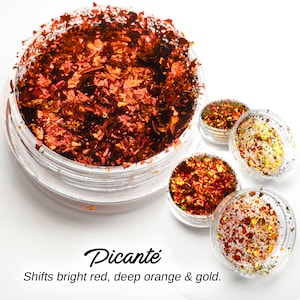May include: A small jar filled with iridescent red, orange, and gold glitter flakes. The jar is labeled "Picante" and the text "Shifts bright red, deep orange & gold." is below the label.