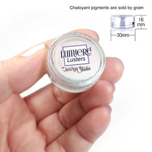May include: A small, clear plastic jar of Lumiere Lusters by JetAge Studio. The jar contains a shimmering, pearlescent pigment. The jar is 30 mm wide and 16 mm high. Text at the top reads "Chatoyant pigments are sold by gram."