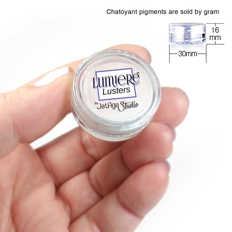 May include: A small, clear plastic jar of Lumiere Lusters by JetAge Studio. The jar contains a shimmering, pearlescent pigment. The jar is 30 mm wide and 16 mm high. Text at the top reads "Chatoyant pigments are sold by gram."