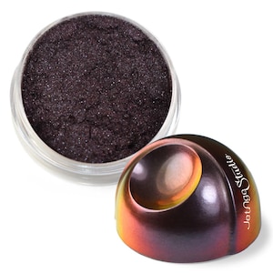May include: A small, round, clear plastic jar filled with dark purple, shimmering cosmetic powder. Beside it sits a dome-shaped lid with a metallic, iridescent finish that shifts between purple, orange, and gold, featuring the text JetAge Studio.
