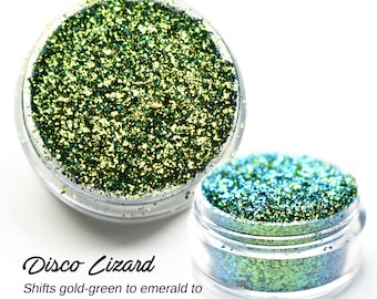 Op de afbeelding: Twee kleine potjes met groene en blauwe, iriserende glitter. De glitter verandert van goudgroen naar smaragdgroen en tealblauw.  "Disco Lizard" staat in cursief onder de potjes.