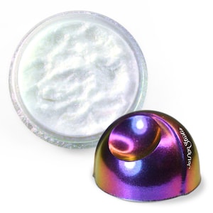 Puede incluir: Un polvo de pigmento blanco iridiscente en un frasco transparente con una muestra de pigmento iridiscente a juego en forma de cúpula. La muestra es de un color vibrante morado, azul y dorado.