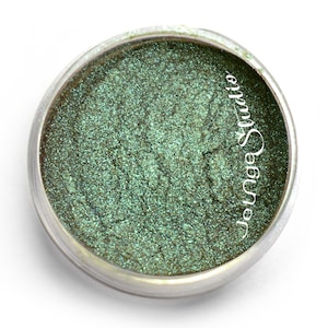 May include: A small, round container of shimmering green eyeshadow. The eyeshadow has a metallic finish and is contained in a clear plastic jar with a silver rim. The text "Jotnge Studio" is printed on the side of the jar.