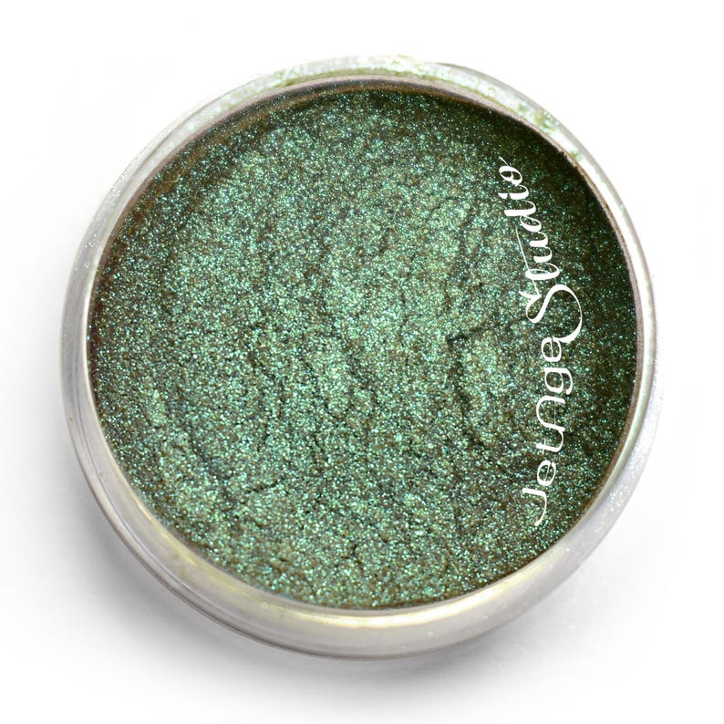 May include: A small, round container of shimmering green eyeshadow. The eyeshadow has a metallic finish and is contained in a clear plastic jar with a silver rim. The text "Jotnge Studio" is printed on the side of the jar.