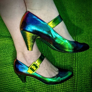 May include: A pair of iridescent green and blue heeled shoes with a buckle strap. The shoes have a high heel and a pointed toe.