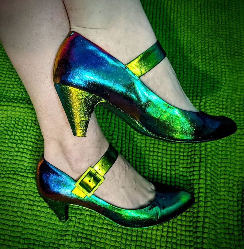 May include: A pair of iridescent green and blue heeled shoes with a buckle strap. The shoes have a high heel and a pointed toe.