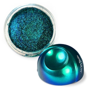 May include: A jar of iridescent teal and blue glitter makeup next to a matching, dome-shaped container. The glitter has a shimmering, granular texture. The container has the text "Lounge Studio" printed on the side.